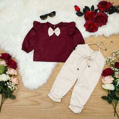 A baby girl toddler outfit with a maroon ruffled shoulder bow tie top and beige elasticated pants.