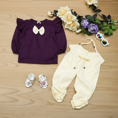 Purple top with a bow and white pants on a wooden surface with flowers and baby shoes.