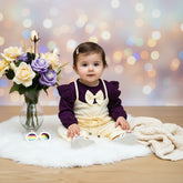 Purple top with ruffled sleeves and white bow, cream pants, baby shoes, and sunglasses on a wooden surface with flowers.