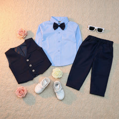 Children's formal outfit with blue shirt, navy pants, vest, shoes, and bow tie on a wooden surface.