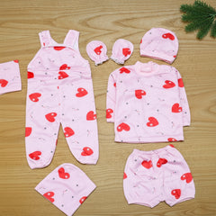 Baby clothing set with pink romper, shirt, pants, and hats with red heart patterns on a wooden surface.