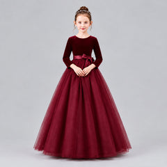 A young girl modeling a formal ball gown with a velvet long-sleeve bodice, a satin waistband, and a voluminous tulle skirt, set against a solid beige studio background.