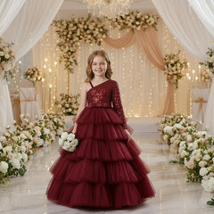 A young girl stands in a decorated event space wearing a formal one-shoulder ball gown with a sparkling upper bodice, a wide silk waistband, and a full layered tulle skirt.