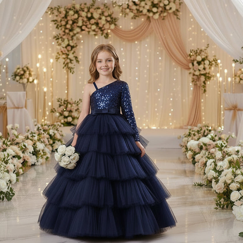A young girl stands in a decorated event space wearing a formal one-shoulder ball gown with a sparkling upper bodice, a wide silk waistband, and a full layered tulle skirt.
