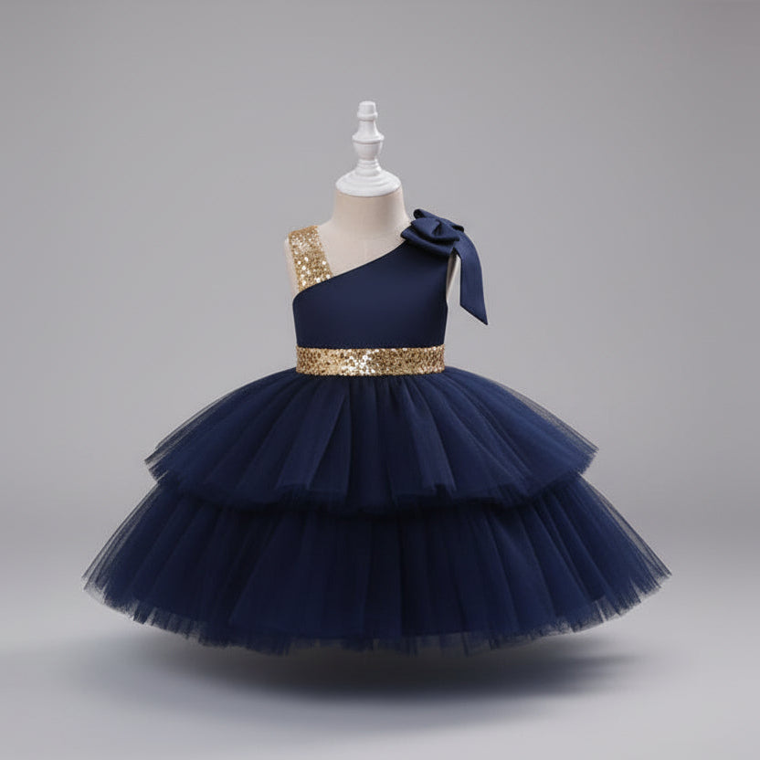 A navy blue one-shoulder toddler party dress with a tiered tulle skirt, a gold sequined waistband, and a matching bow on the shoulder.