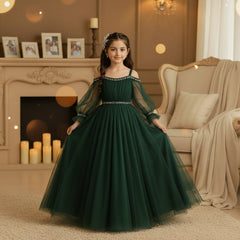 A young girl wearing a bottle green off-the-shoulder tulle ball gown with silver beaded trim at the neckline and waist, standing in a warm beige room.