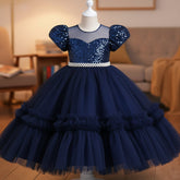 A navy girls' party dress displayed on a mannequin. The dress features a beaded off-the-shoulder bodice with puff sleeves, a pearl-embellished waistband, and a voluminous tiered tulle skirt.