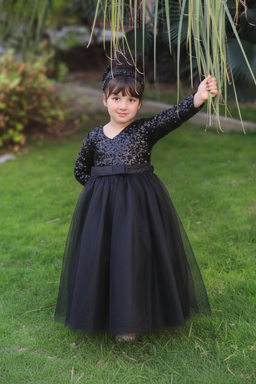 A young girl wearing a long-sleeve ball gown with sequined detailing and a matching headpiece, standing on a green lawn outdoors.