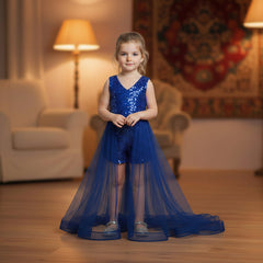 A young girl stands in a warmly lit living room wearing a blue sleeveless lace-trim romper with a long sheer tulle skirt and tulle hem. The room has wooden floors, a cream armchair, two lit floor lamps, and a patterned tapestry on the wall.