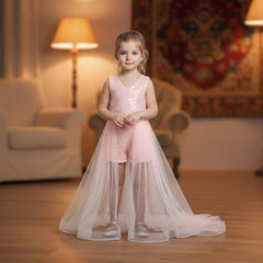 A young girl stands in a warmly lit living room wearing a baby pink sleeveless lace-trim romper with a long sheer tulle skirt and tulle hem. The room has wooden floors, a cream armchair, two lit floor lamps, and a patterned tapestry on the wall.