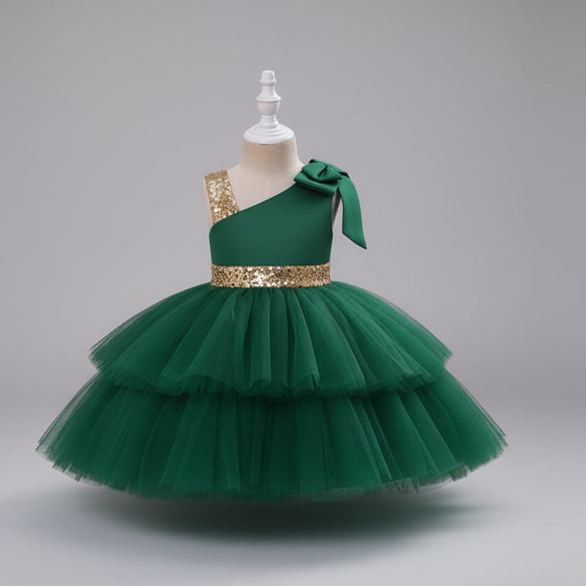 A green one-shoulder toddler party dress with a tiered tulle skirt, a gold sequined waistband, and a matching bow on the shoulder.