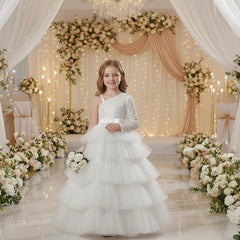 A young girl stands in a decorated event space wearing a formal one-shoulder ball gown with a sparkling upper bodice, a wide silk waistband, and a full layered tulle skirt. 