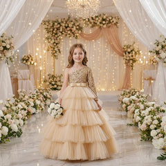 A young girl stands in a decorated event space wearing a formal one-shoulder ball gown with a gold sequined bodice, a wide gold belt, and a full layered tulle skirt.