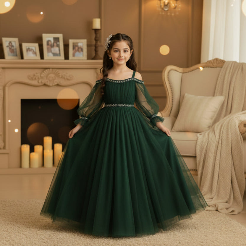 A young girl wearing a bottle green off-the-shoulder tulle ball gown with silver beaded trim at the neckline and waist, standing in a warm beige room.
