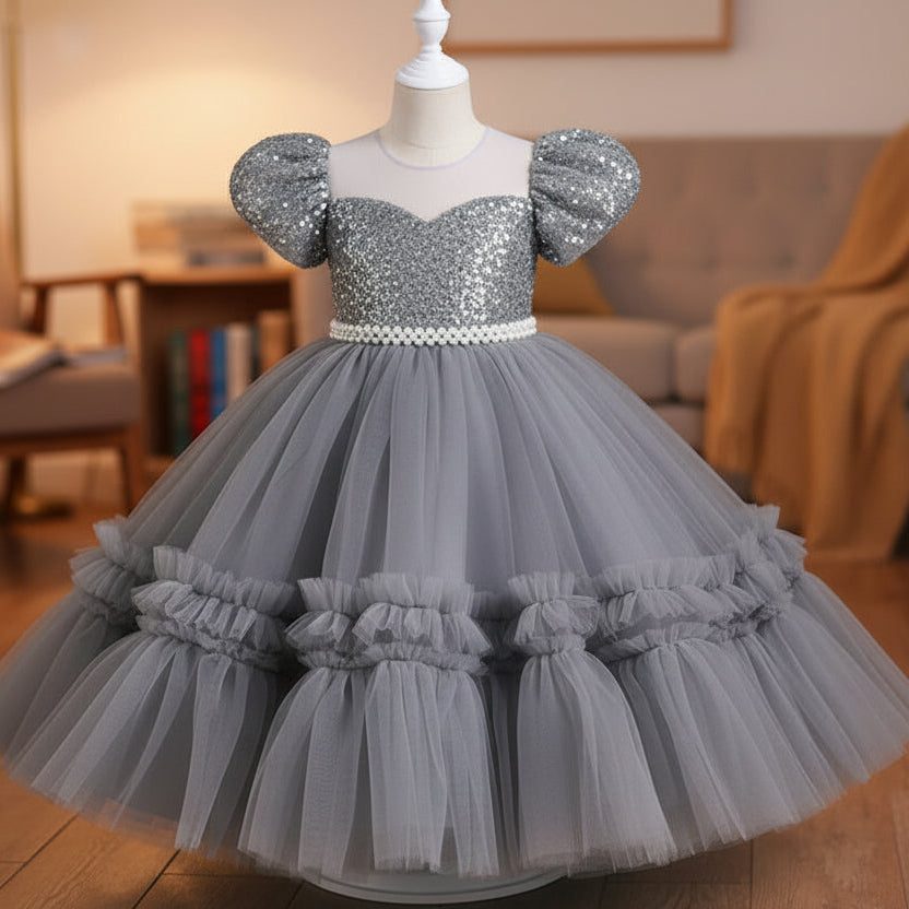 A grey girls' party dress displayed on a mannequin. The dress features a beaded off-the-shoulder bodice with puff sleeves, a pearl-embellished waistband, and a voluminous tiered tulle skirt.