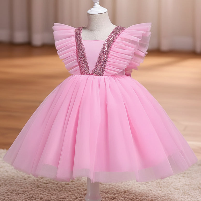 A pink children's party dress displayed on a mannequin against a solid gray background. The dress features a voluminous tulle skirt, ruffled cap sleeves, and a V-neckline embellished with pink sequins.