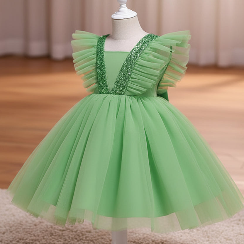 A green children's party dress displayed on a white dress form mannequin. The dress features a voluminous tulle skirt, ruffled cap sleeves, and a V-neckline embellished with sparkling silver sequins, set against a warm indoor background with a wooden floor and beige curtains.