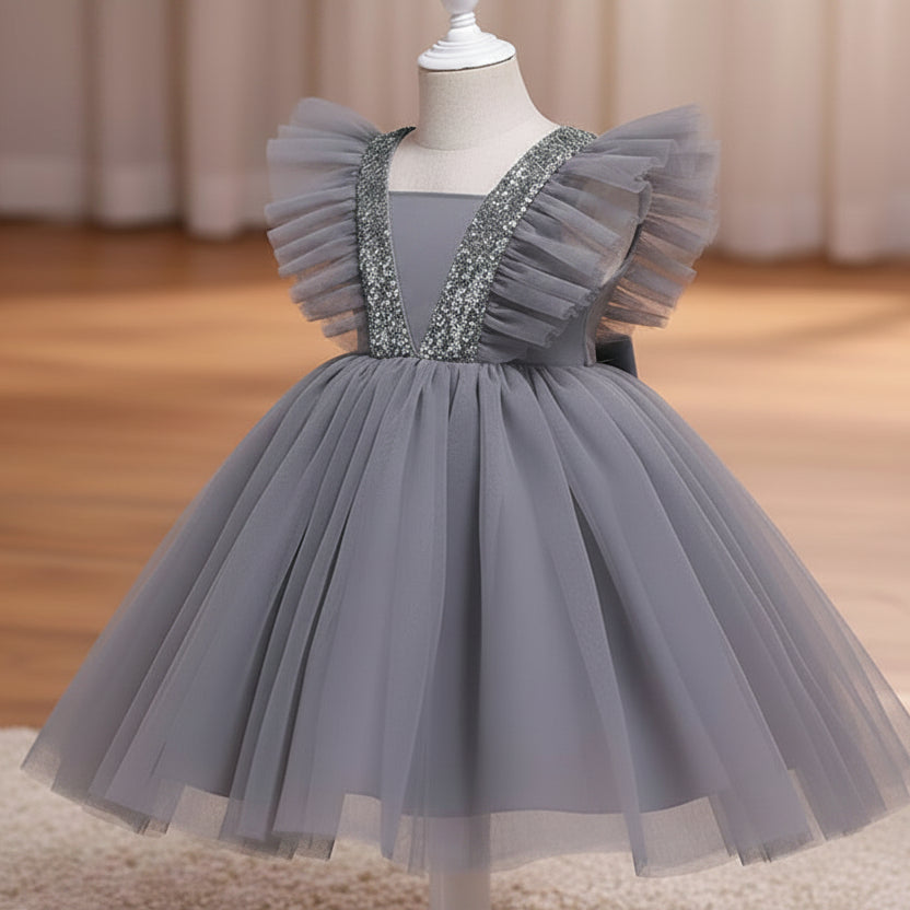 A grey children's party dress displayed on a white dress form mannequin. The dress features a voluminous tulle skirt, ruffled cap sleeves, and a V-neckline embellished with sparkling silver sequins, set against a warm indoor background with a wooden floor and beige curtains.