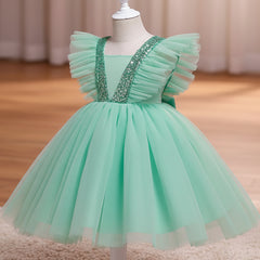 A sea green children's party dress displayed on a white dress form mannequin. The dress features a voluminous tulle skirt, ruffled cap sleeves, and a V-neckline embellished with sparkling silver sequins, set against a warm indoor background with a wooden floor and beige curtains.
