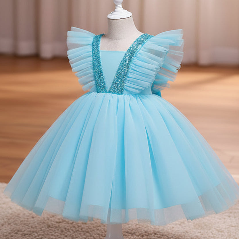 A blue children's party dress displayed on a white dress form mannequin. The dress features a voluminous tulle skirt, ruffled cap sleeves, and a V-neckline embellished with sparkling silver sequins, set against a warm indoor background with a wooden floor and beige curtains.