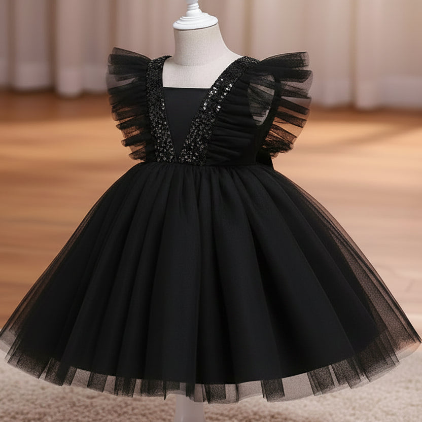 A black children's party dress displayed on a white dress form mannequin. The dress features a voluminous tulle skirt, ruffled cap sleeves, and a V-neckline embellished with sparkling silver sequins, set against a warm indoor background with a wooden floor and beige curtains.