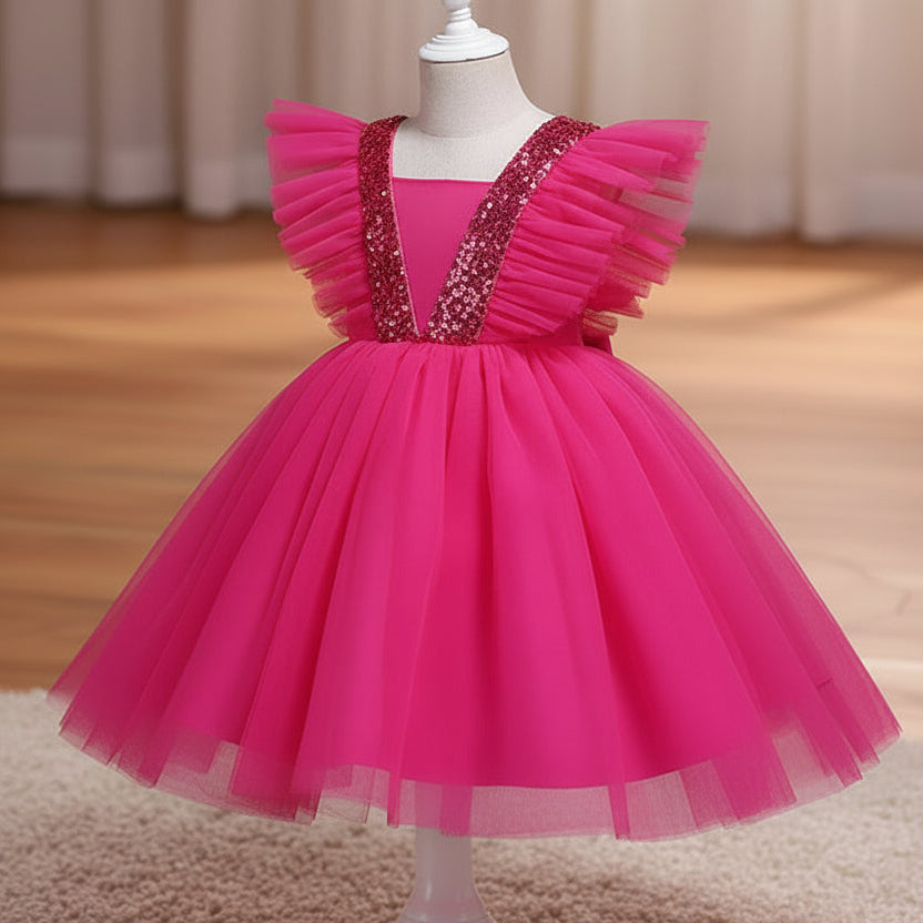A hot pink children's party dress displayed on a white dress form mannequin. The dress features a voluminous tulle skirt, ruffled cap sleeves, and a V-neckline embellished with sparkling silver sequins, set against a warm indoor background with a wooden floor and beige curtains.