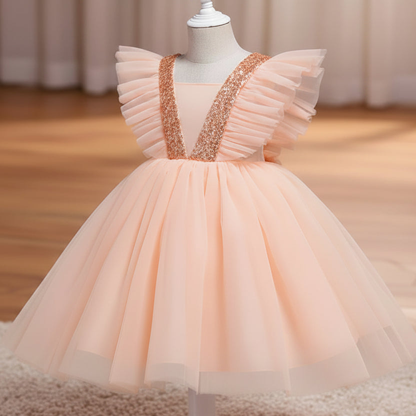 A peach children's party dress displayed on a white dress form mannequin. The dress features a voluminous tulle skirt, ruffled cap sleeves, and a V-neckline embellished with sparkling silver sequins, set against a warm indoor background with a wooden floor and beige curtains.