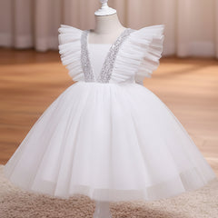 A white children's party dress displayed on a white dress form mannequin. The dress features a voluminous tulle skirt, ruffled cap sleeves, and a V-neckline embellished with sparkling silver sequins, set against a warm indoor background with a wooden floor and beige curtains.