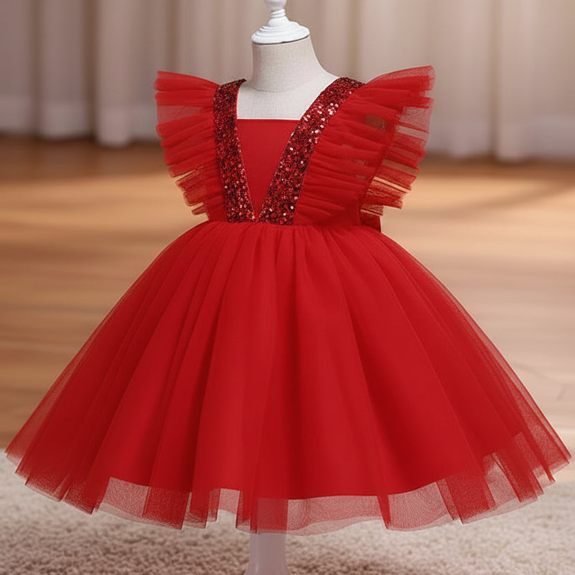 A red children's party dress displayed on a white dress form mannequin. The dress features a voluminous tulle skirt, ruffled cap sleeves, and a V-neckline embellished with sparkling silver sequins, set against a warm indoor background with a wooden floor and beige curtains.