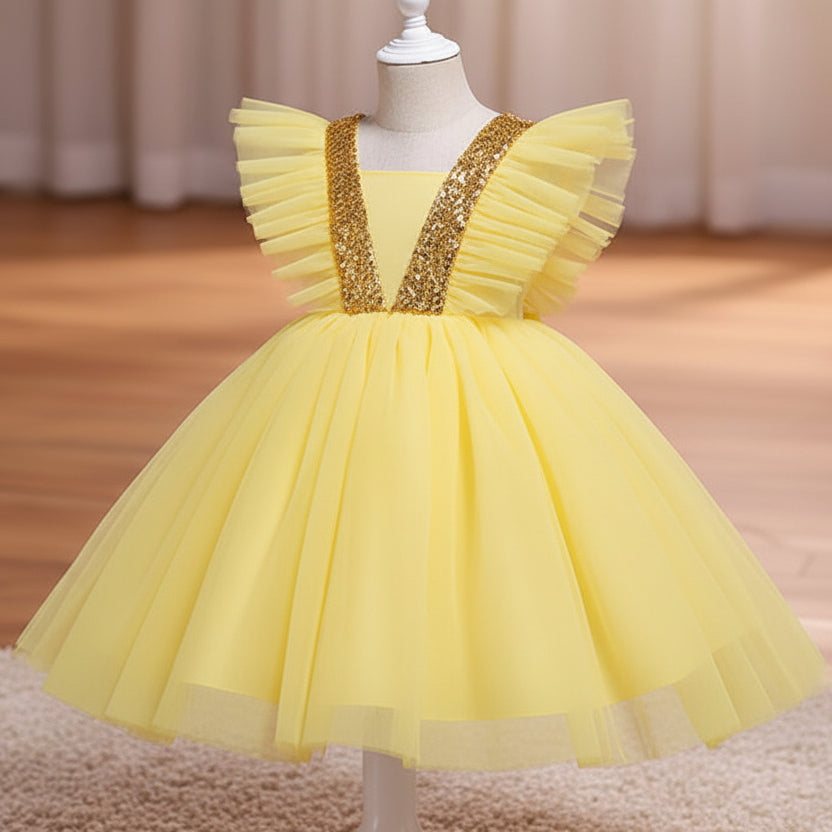 A yellow children's party dress displayed on a white dress form mannequin. The dress features a voluminous tulle skirt, ruffled cap sleeves, and a V-neckline embellished with sparkling silver sequins, set against a warm indoor background with a wooden floor and beige curtains.