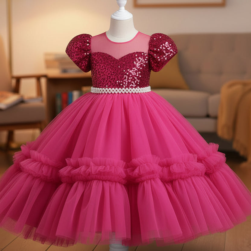 A shocking pink sequined girl's party dress with puff sleeves and a tiered tulle skirt, displayed on a white mannequin in a cozy living room setting.