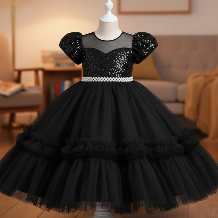 A black sequined girl's party dress with puff sleeves and a tiered tulle skirt, displayed on a white mannequin in a cozy living room setting.