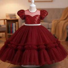 A maroon girls' party dress displayed on a mannequin. The dress features a beaded off-the-shoulder bodice with puff sleeves, a pearl-embellished waistband, and a voluminous tiered tulle skirt.