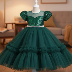 A sea green sequined girl's party dress with puff sleeves and a tiered tulle skirt, displayed on a white mannequin in a cozy living room setting.
