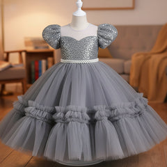 A grey girls' party dress displayed on a mannequin. The dress features a beaded off-the-shoulder bodice with puff sleeves, a pearl-embellished waistband, and a voluminous tiered tulle skirt.