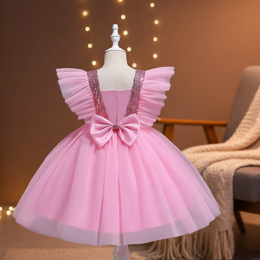 A pink children's party dress displayed on a mannequin, featuring a voluminous tulle skirt, a large bow at the back, and ruffled cap sleeves with sequined shoulder panels.