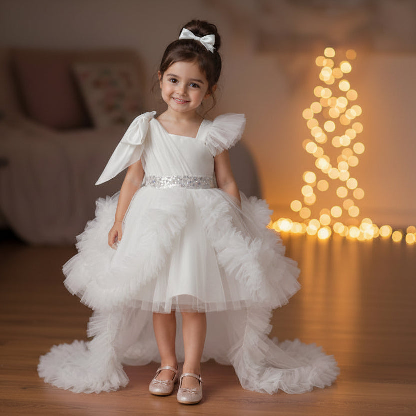 A young girl stands in a warmly lit living room wearing a white party dress with a high-low ruffled skirt, a bow headband, and metallic shoes. 