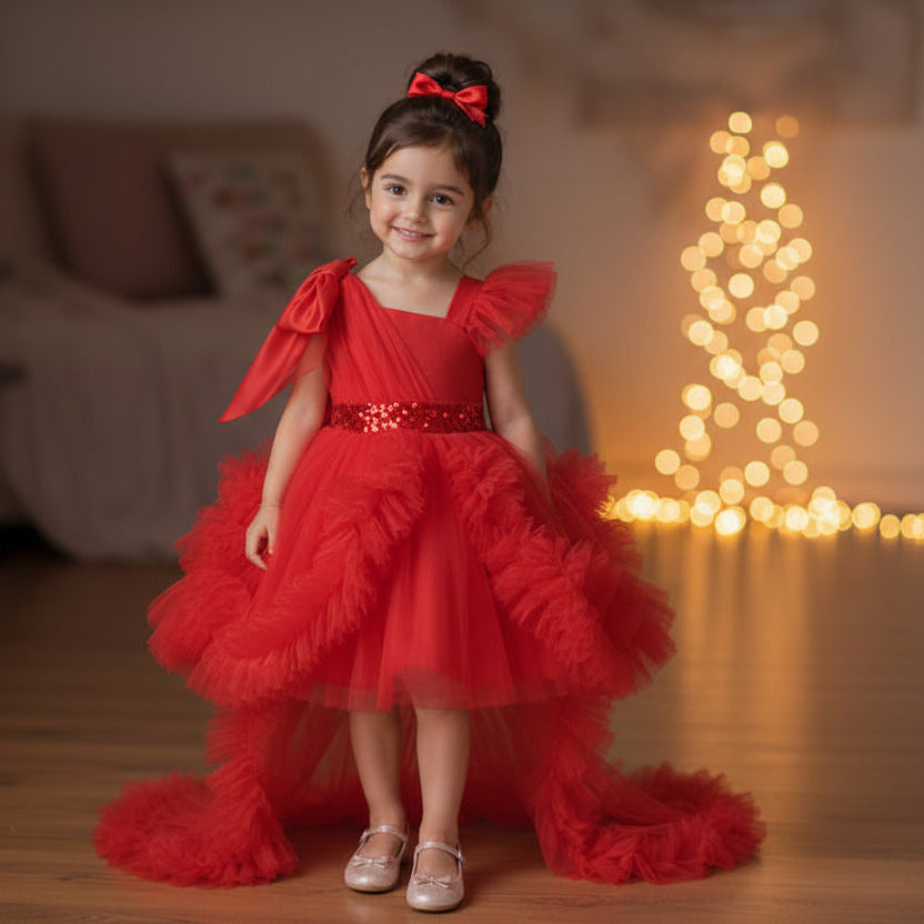 A young girl stands in a warmly lit living room wearing a red party dress with a high-low ruffled skirt, a bow headband, and metallic shoes. 