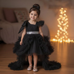 A young girl stands in a warmly lit living room wearing a black party dress with a high-low ruffled skirt, a bow headband, and metallic shoes.