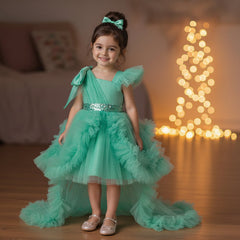 A young girl stands in a warmly lit living room wearing a sea green party dress with a high-low ruffled skirt, a bow headband, and metallic shoes. 